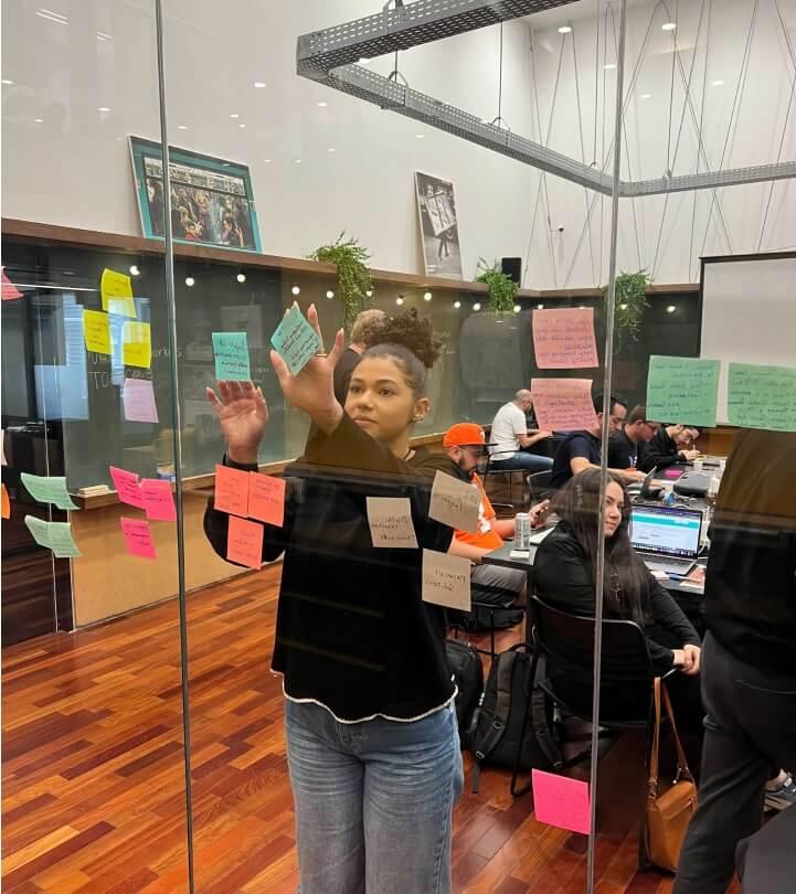 Photo of a young woman at Able in a team meeting contributing ideas by adding notes to a glass conference room wall